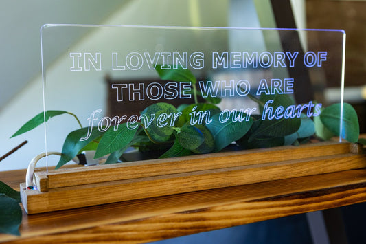 Image depicts LED lamp with oak wood base and scratch-resistant acrylic engraved with the phrase "in loving memory of those forever in our hearts" and displayed on a wooden shelf with greenery. The lamp is used as wedding decor to memorialize loved ones no longer with us.
